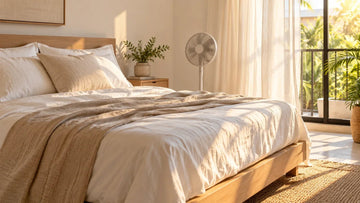 Cama con sábanas claras y ventilador junto a una ventana abierta en clima cálido